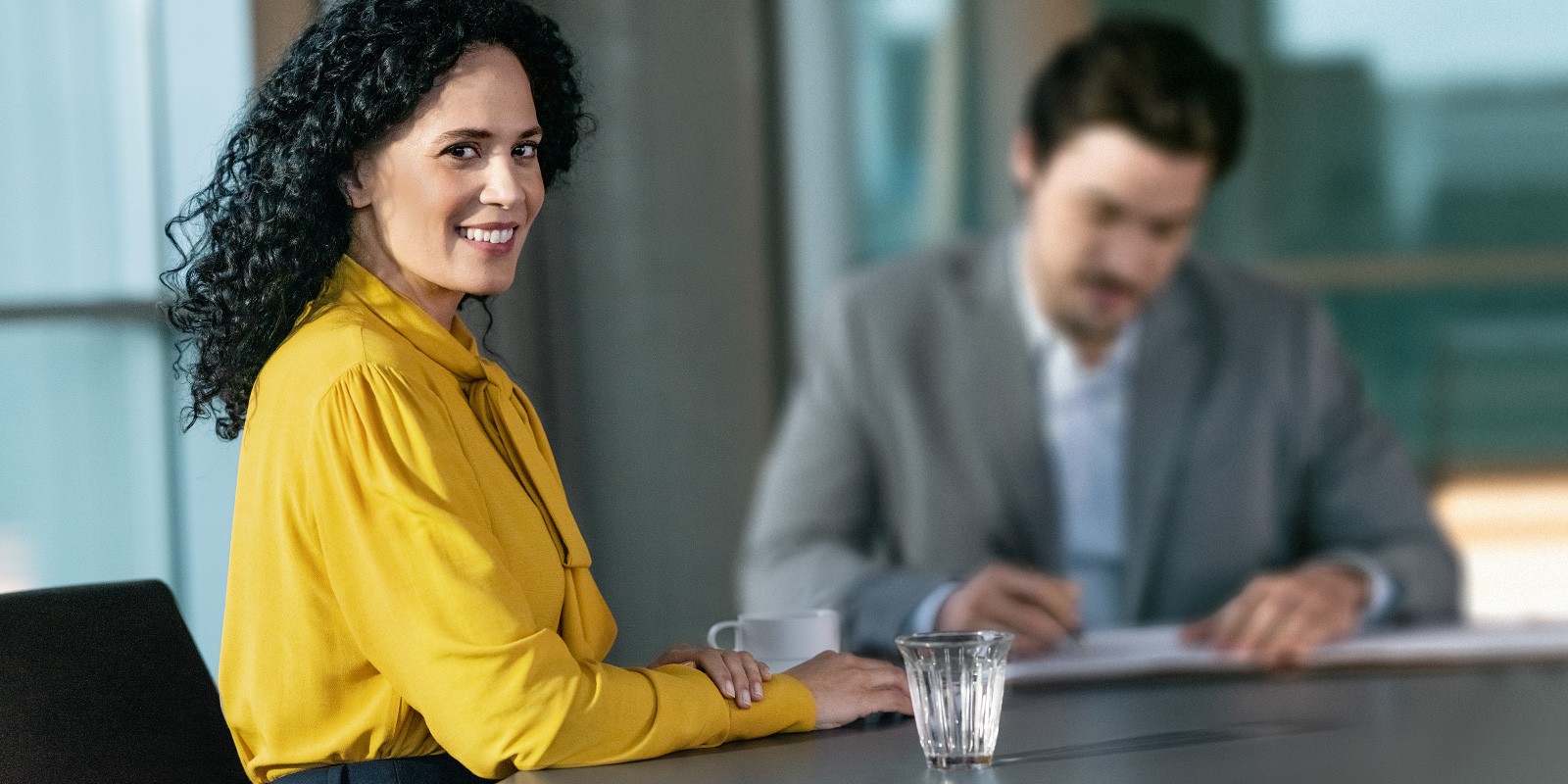 Das Bild zeigt eine Frau mit einer gelben Bluse, die lächelnd direkt in die Kamera schaut und links neben ihrem Steuerberater an einem Schreibtisch sitzt. 