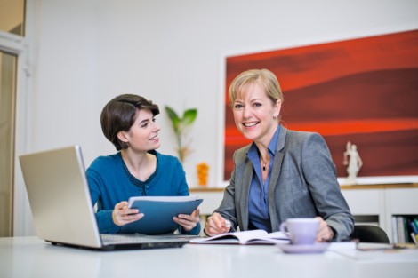 zwei Frauen am Schreibtisch, vor ihnen ein Laptop. Im Hintergrund ein rotes Bild